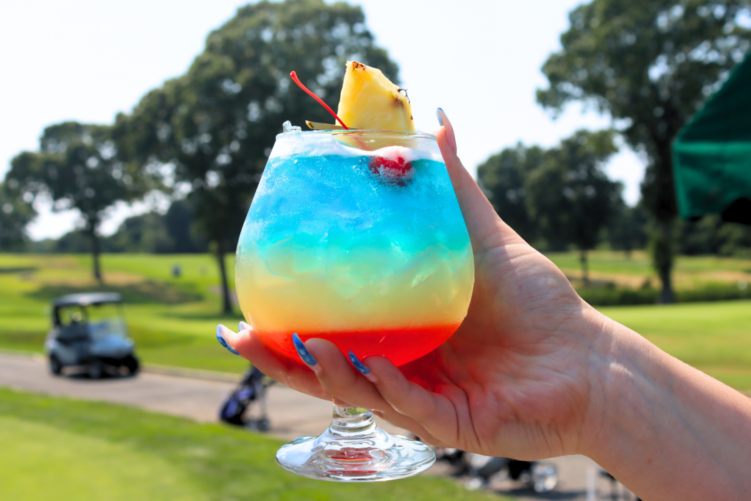 A hand holding a colorful layered cocktail garnished with a pineapple slice and cherry, with a golf course in the background.