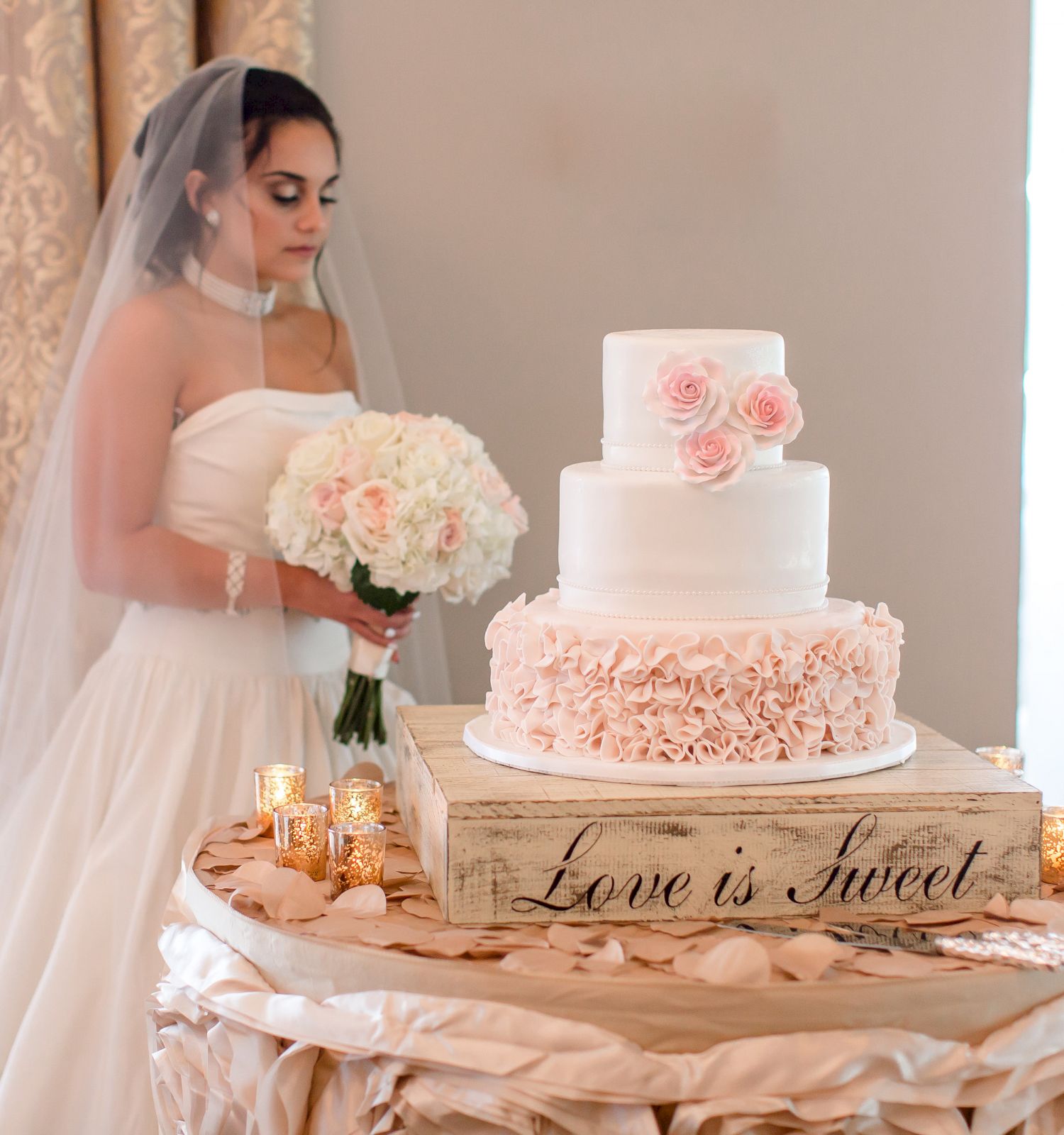 A bride, holding a bouquet, stands next to a white and pink wedding cake on a decorated table with a sign that reads 