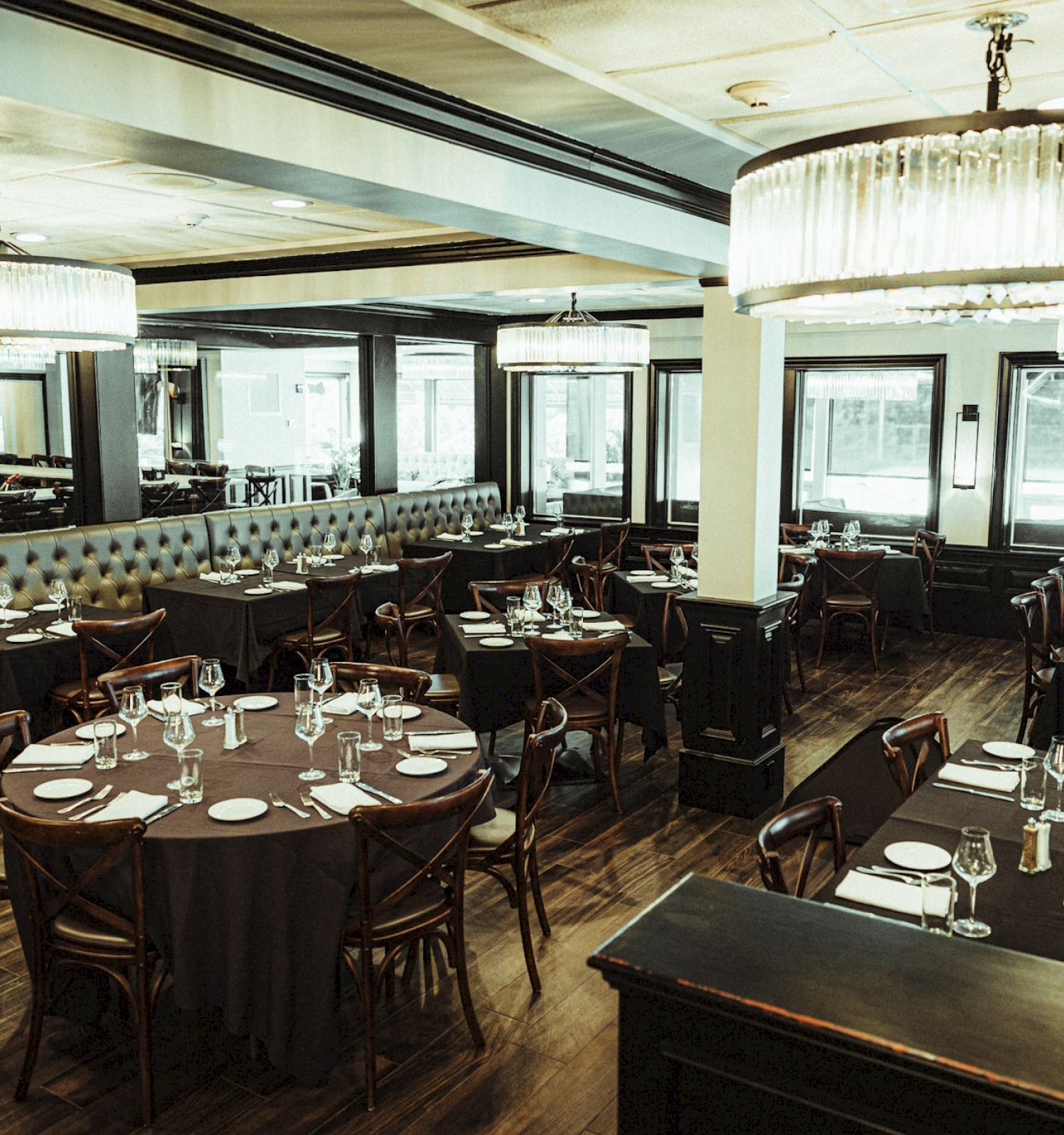 The image shows a classy restaurant interior with round tables set with plates and glassware, elegant chandeliers, and large windows.