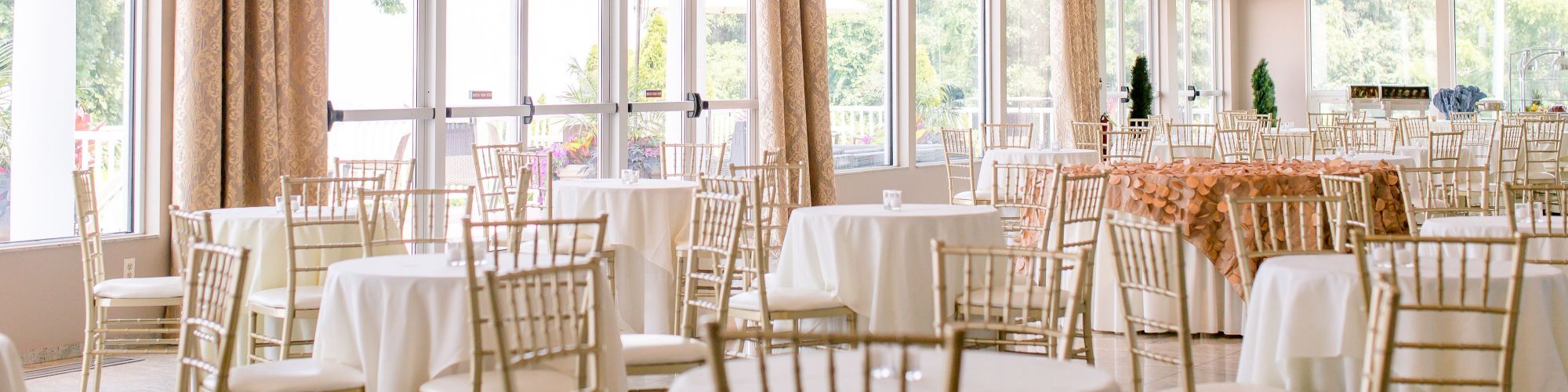 An elegant dining hall with white tablecloth-covered round tables, gold chairs, large windows, and natural light streaming in, appears set for an event.