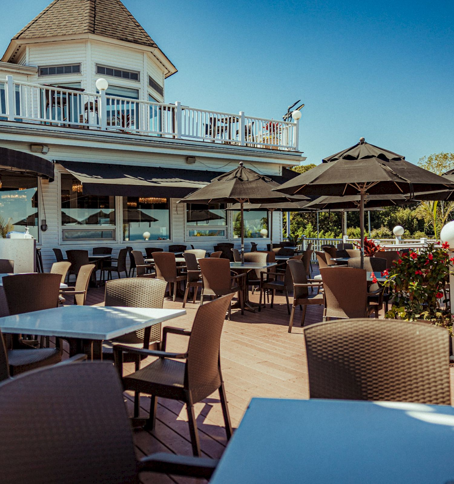 An outdoor dining area with tables and chairs, shaded by black umbrellas, near a two-story building with a balcony.