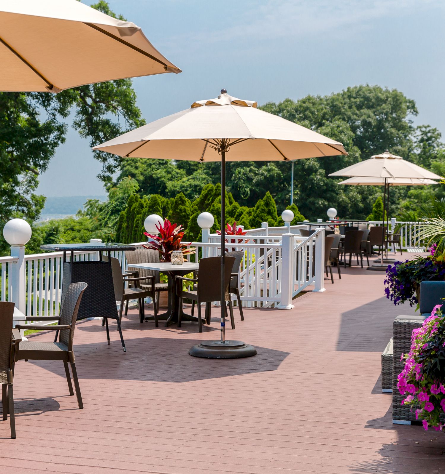 An outdoor patio area with tables, chairs, and umbrellas, surrounded by plants and flowers, overlooking a scenic view.