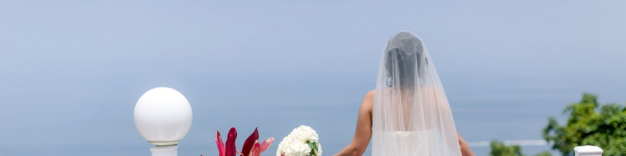 A bride in a wedding dress and veil stands on a balcony, holding a bouquet and looking out at a scenic, calm ocean view.