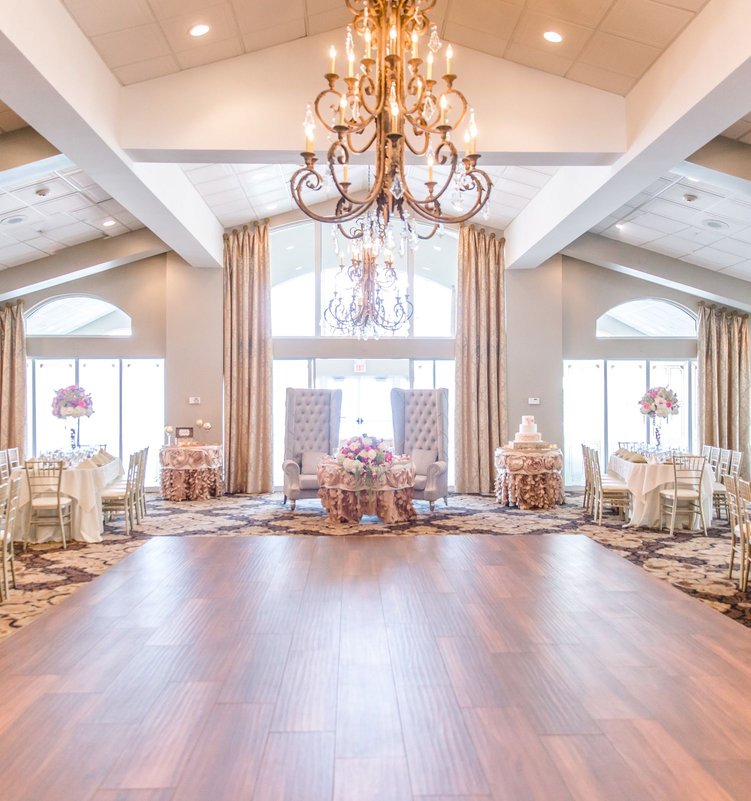 This image depicts an elegant ballroom set for an event with chandeliers, round tables with floral centerpieces, and a polished wooden floor.