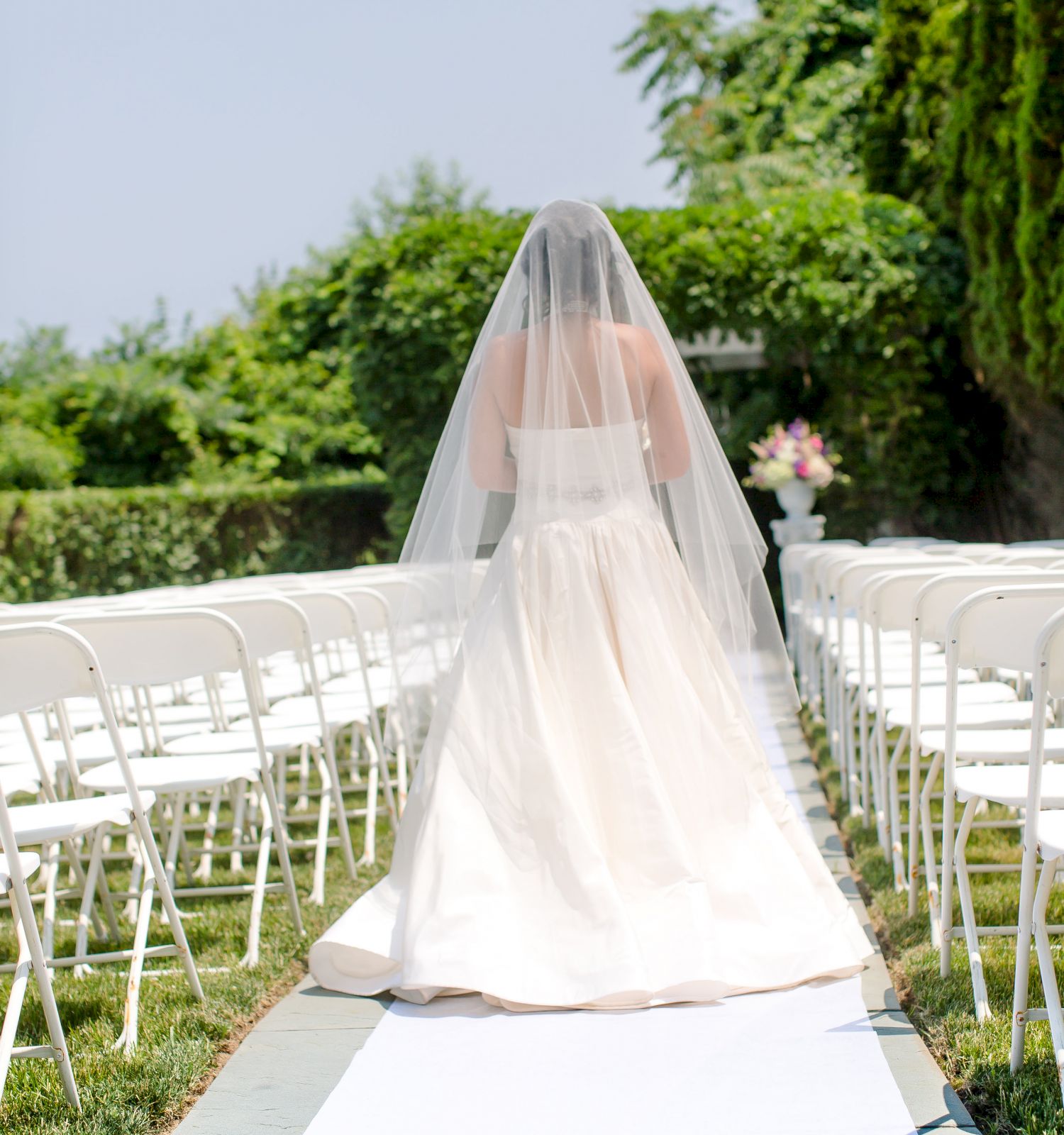 A bride with a veil walks down an outdoor aisle lined with white chairs amongst greenery, on a sunny day.