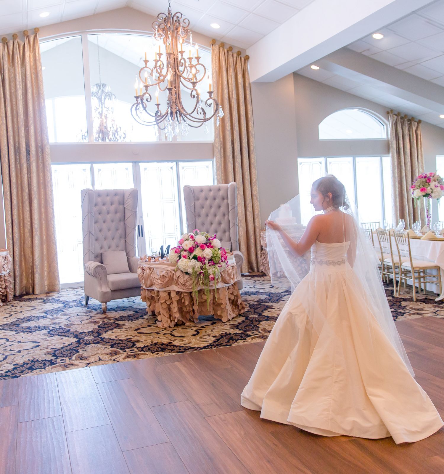 A woman in a wedding dress stands on a wooden floor in a decorated, elegant reception hall with chandeliers, flowers, and seated areas.