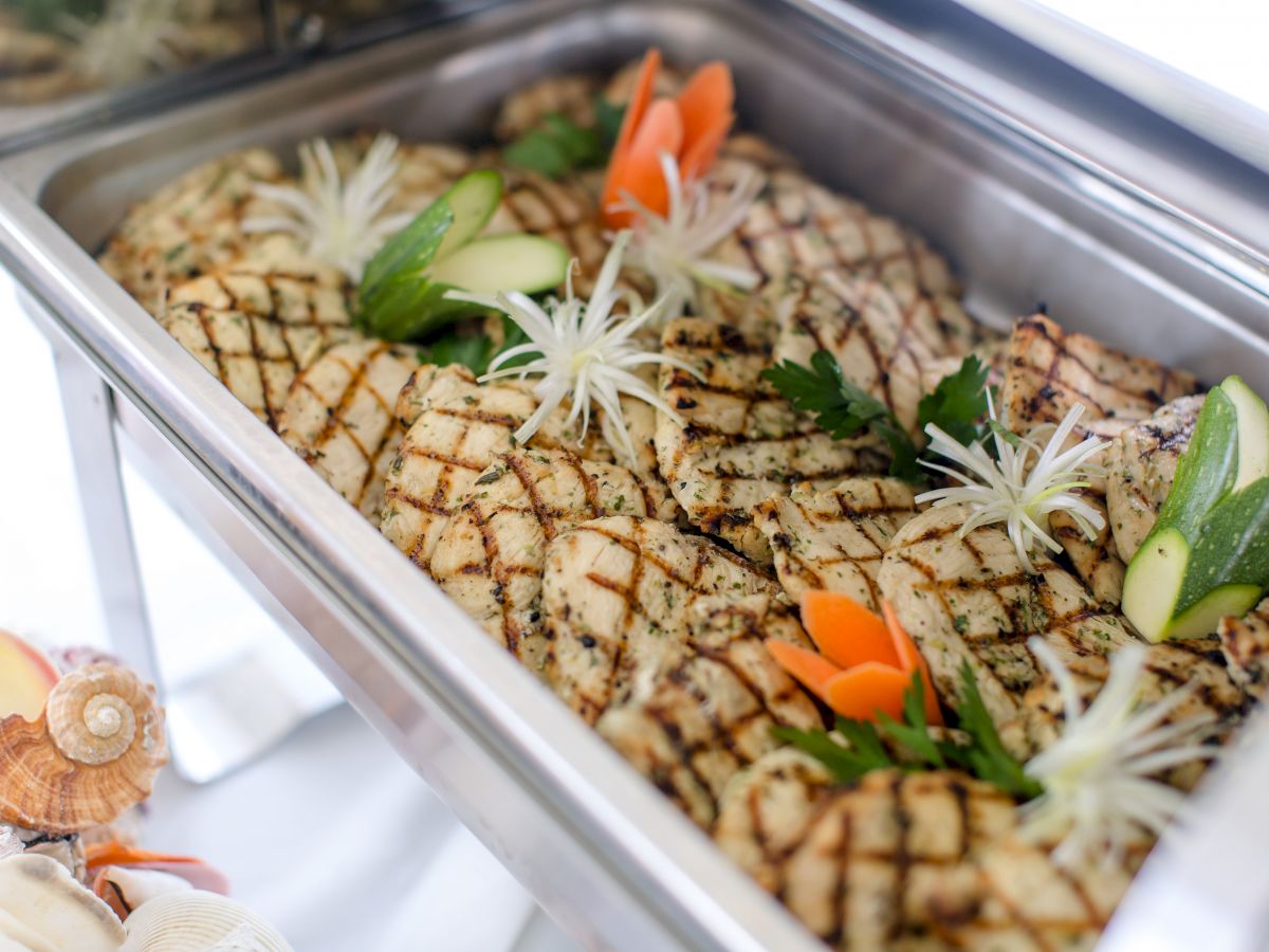 A chafing dish filled with grilled, seasoned fish, garnished with slices of zucchini, fresh parsley, and artfully cut vegetables.