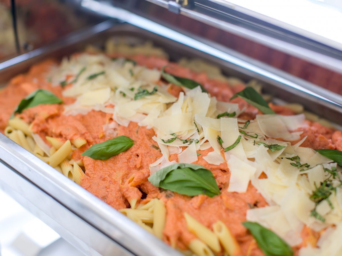 A buffet tray filled with penne pasta topped with creamy tomato sauce, basil leaves, and shaved Parmesan cheese.