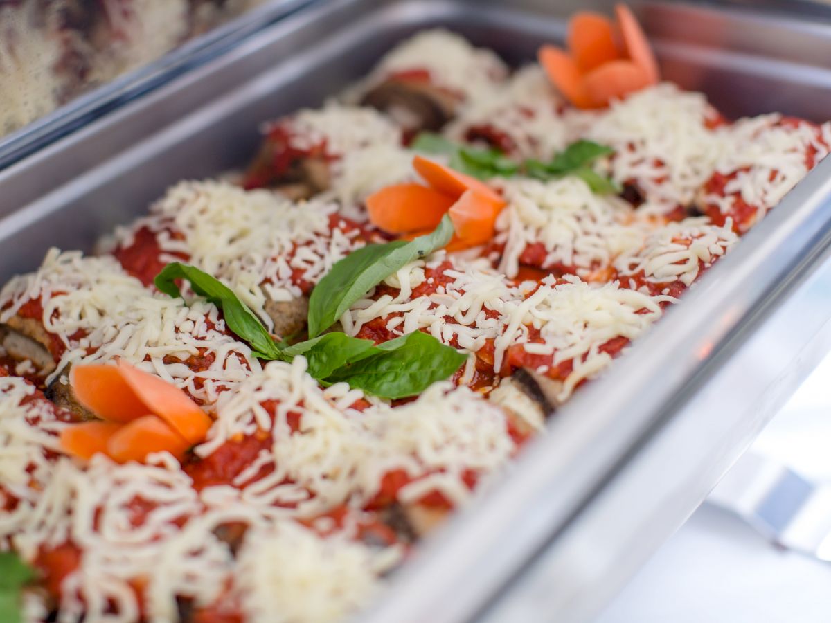 The image shows a tray of baked dish topped with melted cheese, tomato sauce, and garnished with fresh basil and carrot slices.