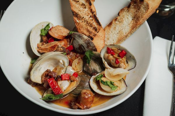 A plate of clams with red peppers and herbs, served with two pieces of toasted bread, next to a white napkin with a fork.