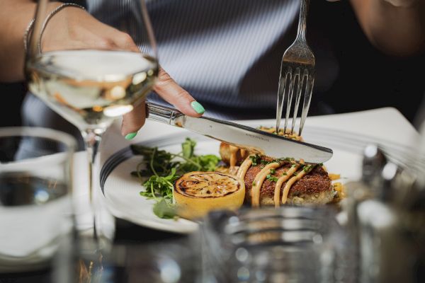 A person is cutting food with a knife and fork, surrounded by a glass of wine, lemon slice, and greens on a white plate.