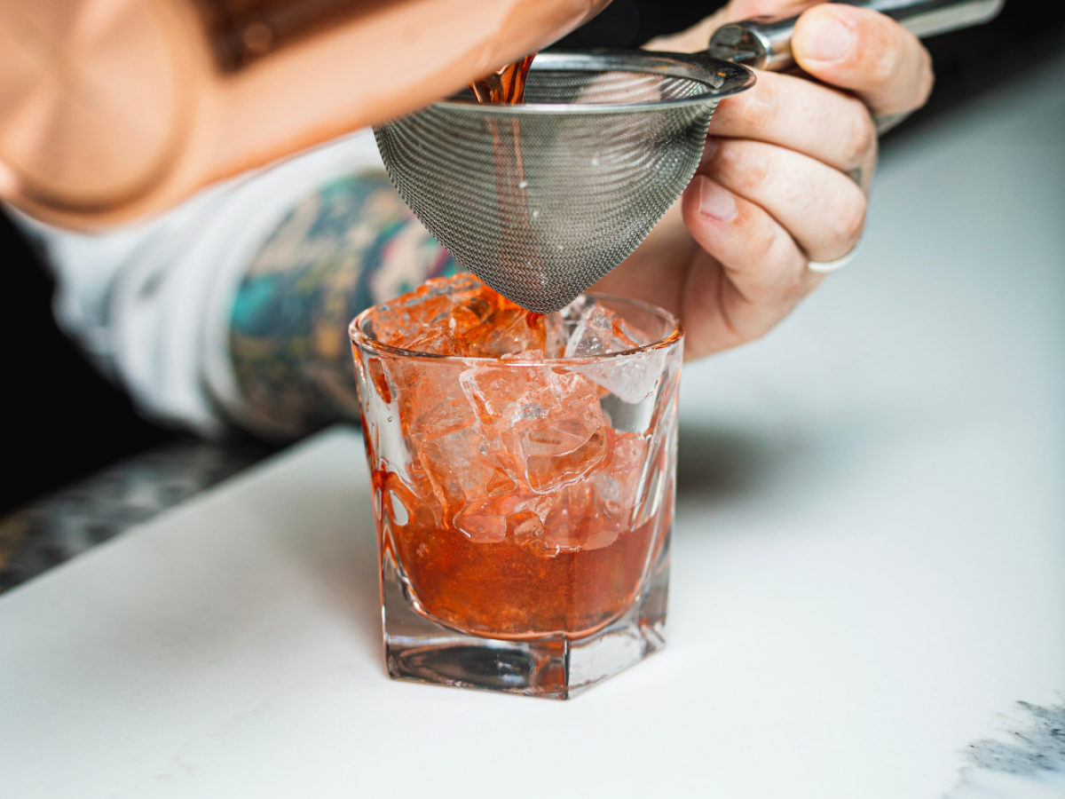 A person is pouring a red liquid through a strainer into a glass filled with ice, using a copper cocktail shaker on a counter, always ending the sentence.