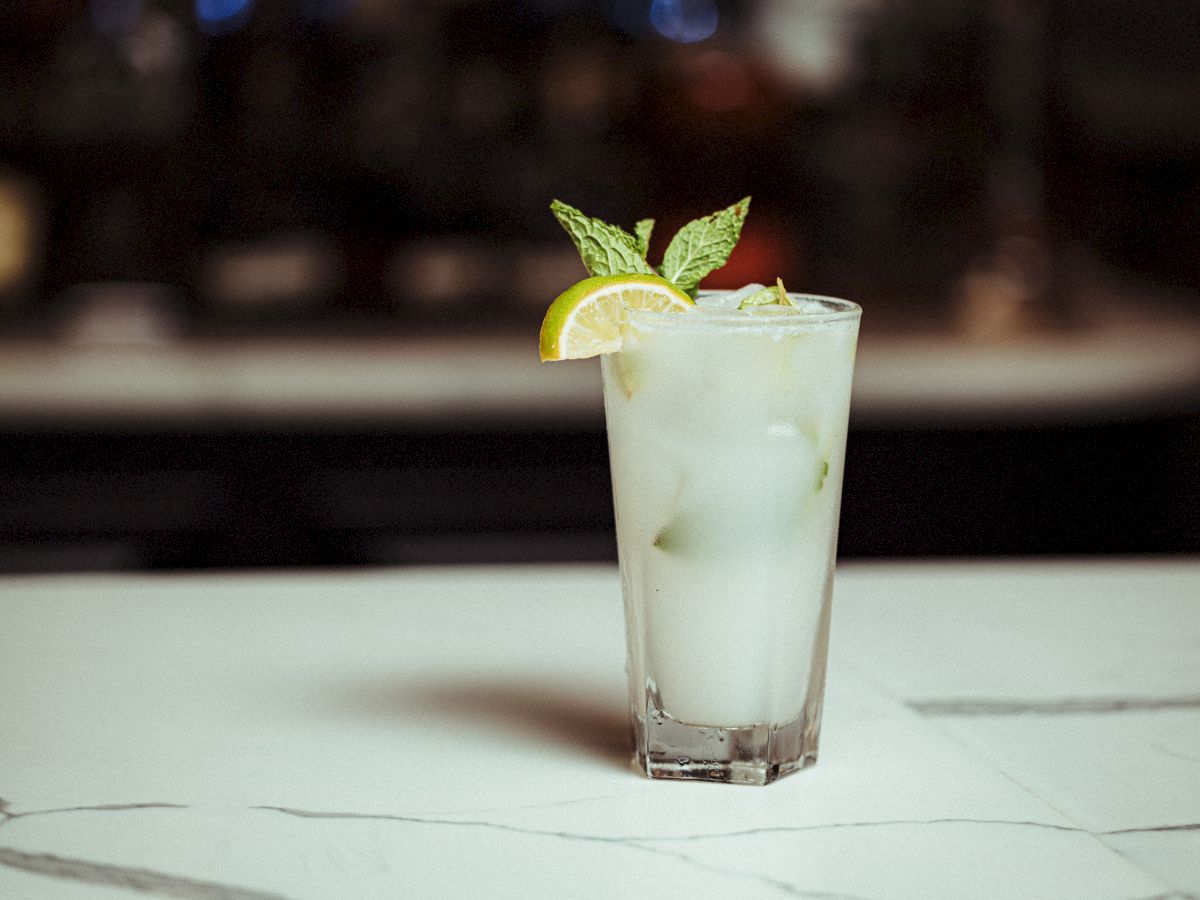 A tall glass filled with an iced beverage, garnished with a lemon wedge and mint leaves, sitting on a marble countertop.