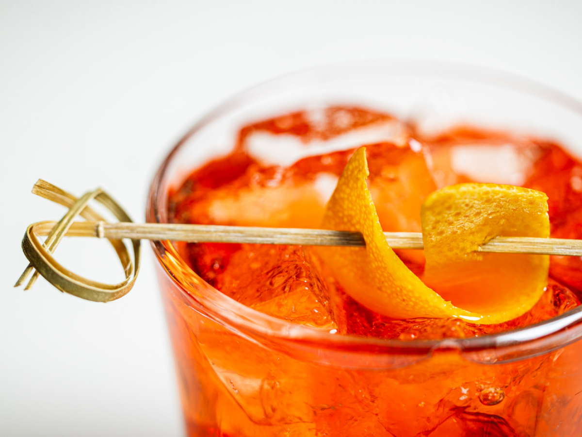 A glass filled with an orange-colored beverage, garnished with an orange peel on a skewer, resting on ice cubes.