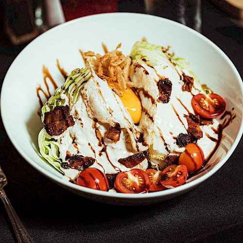 A bowl with sliced lettuce, cherry tomatoes, poached egg, bacon bits, and sauce, garnished with fried onions. Forks and a glass are beside it.