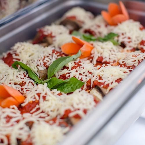 A stainless steel chafing dish filled with a layered dish topped with melted cheese, tomato sauce, basil, and garnished with orange vegetables.