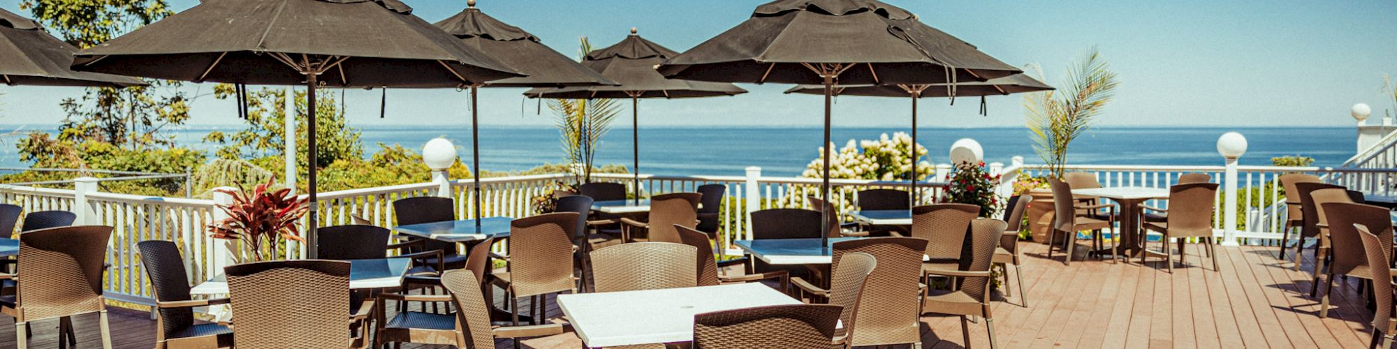 An outdoor dining area with multiple tables and chairs, shaded by black umbrellas, overlooking a scenic ocean view under a clear, blue sky.