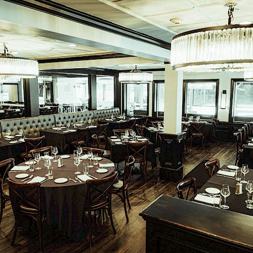 The image shows an elegant, empty restaurant with round and rectangular tables set with white plates and glassware, under large chandeliers.