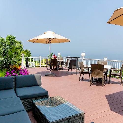 An outdoor deck with seating, umbrellas, and a sea view, surrounded by a white railing, with plants and flowers adding colorful touches.