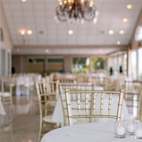 A beautifully decorated event hall with gold chairs and round tables covered in white tablecloths, candles on tables and a chandelier above.