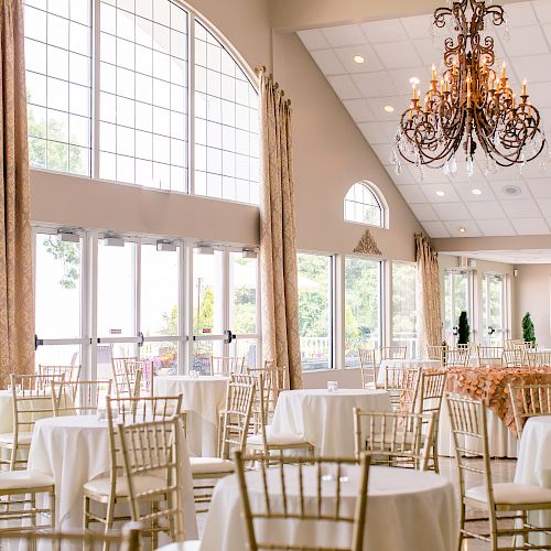 A spacious, elegant event hall with round tables covered in white cloth, golden chairs, large windows, and a chandelier hanging from the ceiling.