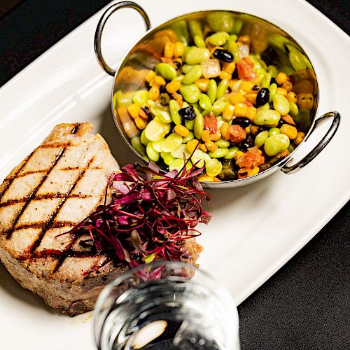 The image shows a grilled steak on a white plate, garnished with microgreens, alongside a small bowl of a colorful vegetable medley.