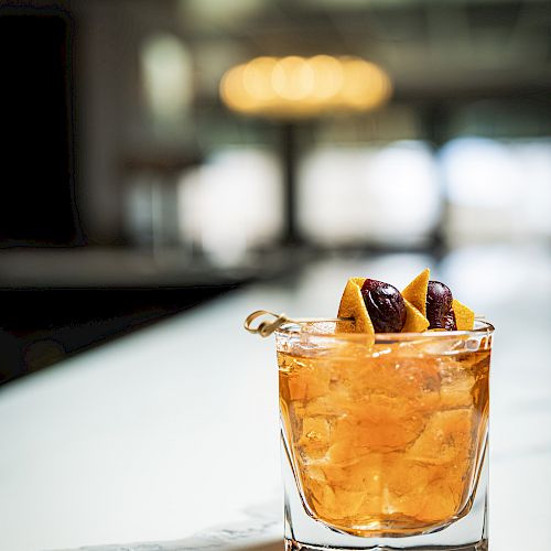 A glass of an amber-colored cocktail, garnished with orange twists and dark cherries on a skewer, is placed on a white surface in a softly-lit setting.