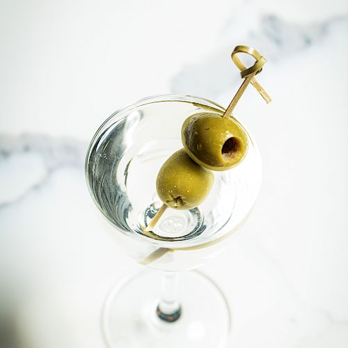 A martini glass filled with a clear liquid, garnished with two green olives on a cocktail pick.