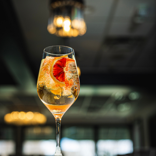 A cocktail glass with a drink garnished with an orange slice and ice cubes, set against the backdrop of a dimly lit indoor setting.