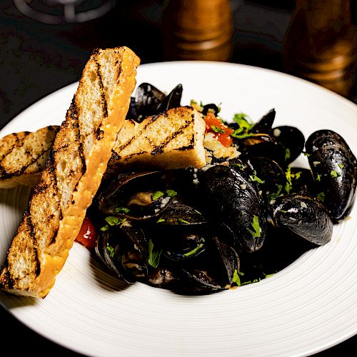 A dish of mussels garnished with herbs and tomatoes, served with two grilled bread slices on a white plate, with a drink and condiments nearby.