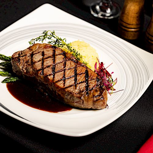 A grilled steak garnished with herbs served on a plate with vegetables and a side of mashed potatoes. There’s also a glass of water and pepper mills.