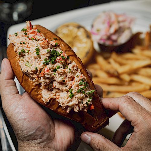 A person holds a lobster roll, garnished with herbs, with a side of fries and coleslaw on the plate in the background.