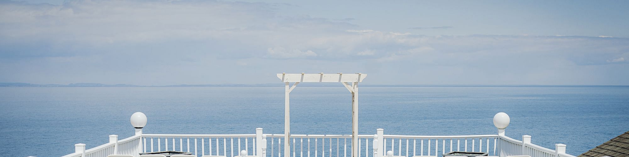 An outdoor wedding setup with white chairs arranged on either side of an aisle, leading to an arch overlooking the ocean, with flower arrangements.