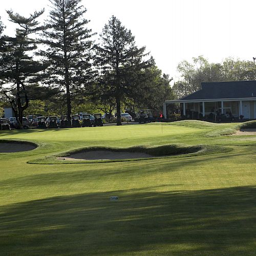 A manicured golf course with sand traps, surrounded by trees, and a clubhouse in the background; golf carts are lined up near the clubhouse.