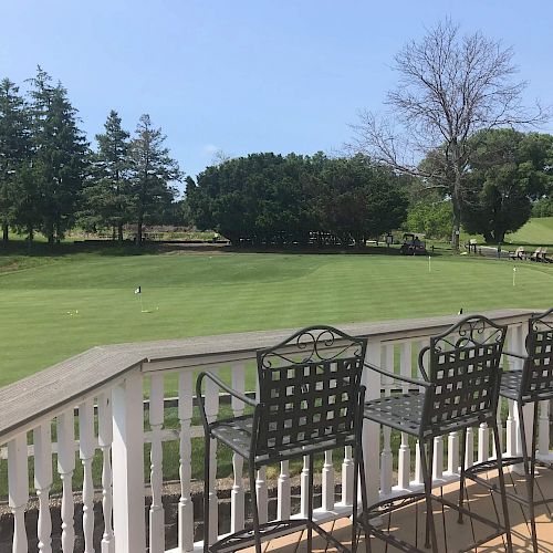 A deck with three high chairs overlooks a green golf course surrounded by trees. The sky is clear and blue, and the setting is serene.