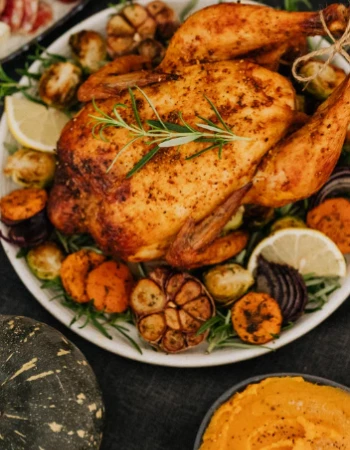 A roasted chicken garnished with herbs, lemon, and vegetables. Accompanied by a bowl of mashed squash and assorted sides.