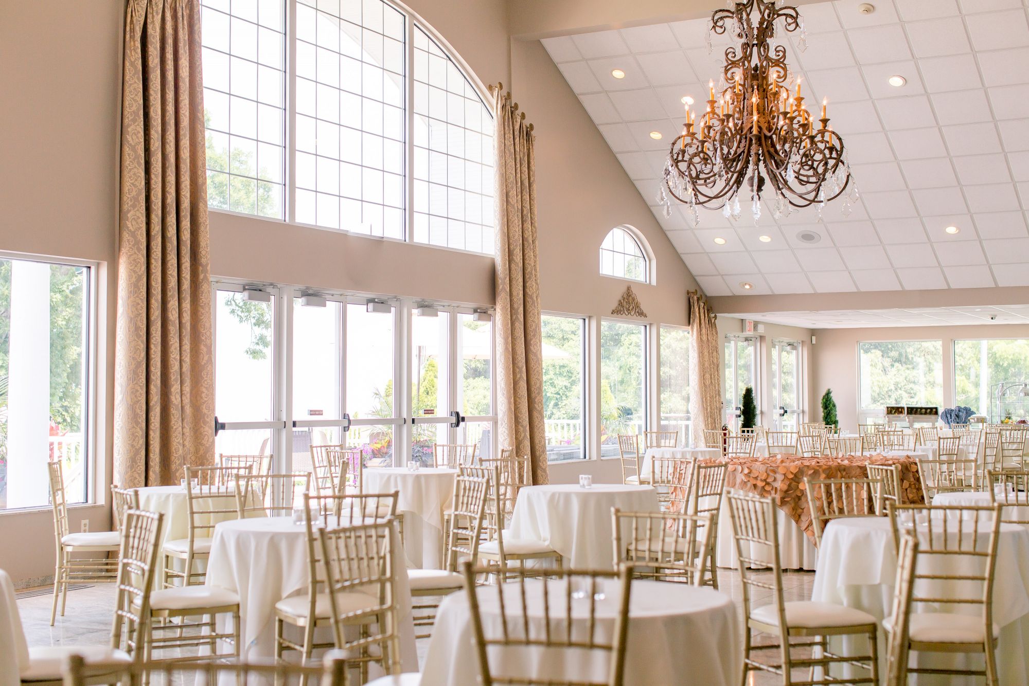 An elegant banquet hall is set up, featuring round tables with white tablecloths, gold chairs, and a large chandelier.