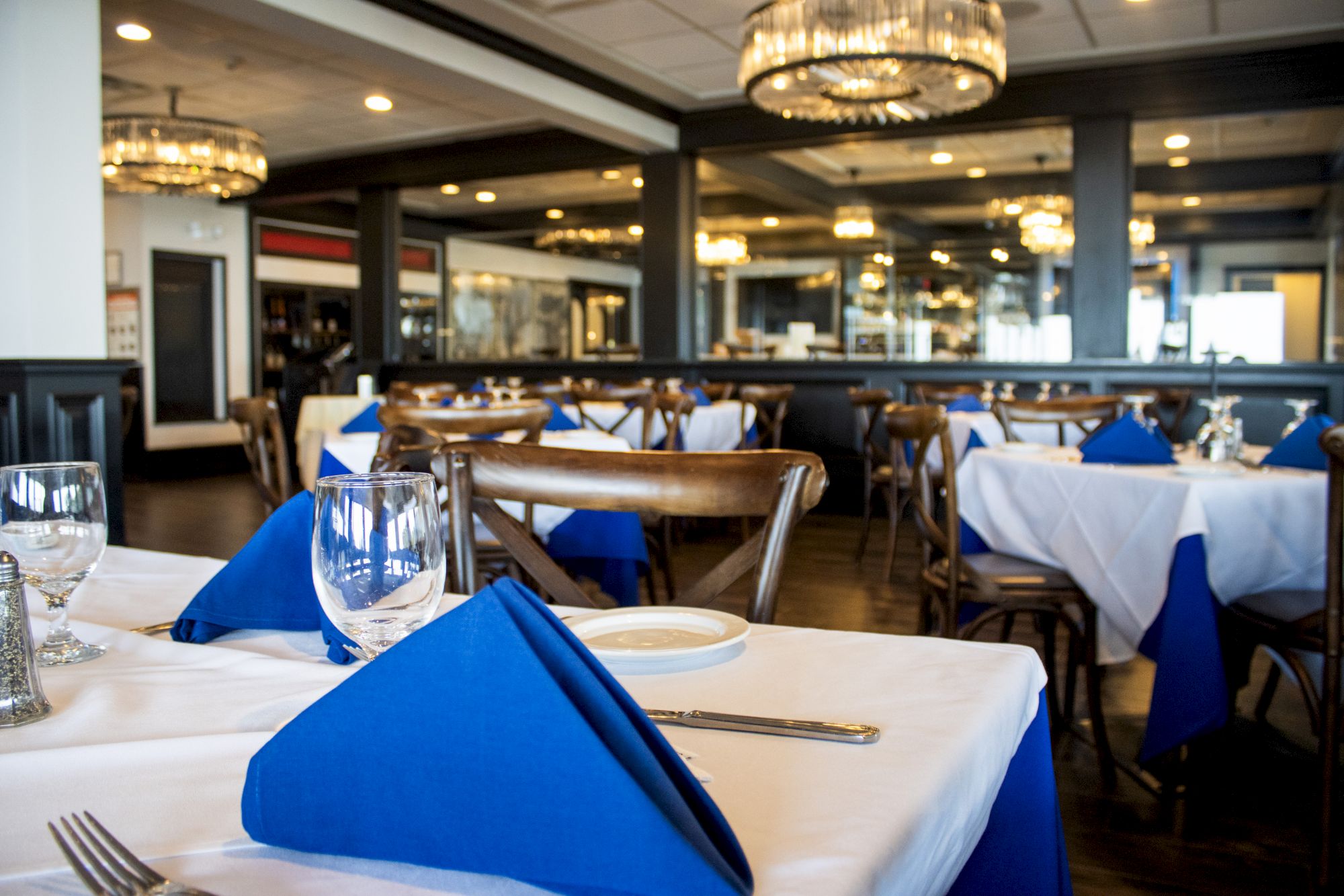 The image shows a restaurant interior with tables set with white cloths and blue napkins, surrounded by wooden chairs and chandeliers.