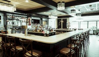 A modern bar with luxurious chandeliers, a marble countertop, stocked shelves, and wooden barstools in an elegant setting with ample lighting.