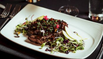 A gourmet dish on a white plate features seared meat topped with herbs and edible flowers, with a side of greens and apple slices.