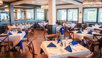 A restaurant dining area with neatly arranged tables, white tablecloths, blue napkins, and wooden chairs, under elegant pendant lights.