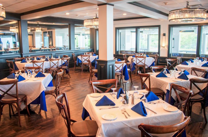 A restaurant dining area with neatly arranged tables, white tablecloths, blue napkins, and wooden chairs, under elegant pendant lights.