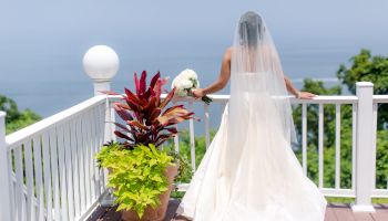 A bride in a white dress and veil stands on a balcony overlooking the ocean, holding a bouquet of flowers and next to a potted plant with red leaves.