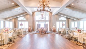 A spacious, elegant banquet hall set for an event, with round tables, gold chairs, floral centerpieces, and chandeliers hanging from a vaulted ceiling.