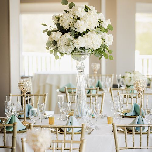 An elegantly decorated event space with white floral centerpieces, gold chairs, neatly arranged table settings, and folded napkins.