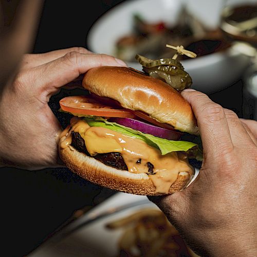 A person is holding a hamburger with lettuce, tomato, onion, and melted cheese, with fries and a drink in the background.