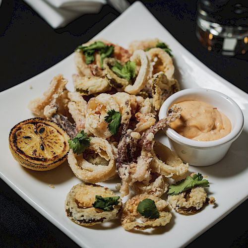 A plate of fried calamari with a lemon wedge and a small bowl of dipping sauce, garnished with green herbs, on a white square dish.