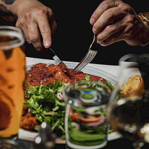 A person is using a knife and fork to eat a meal, which includes a breaded dish and a fresh salad, accompanied by a glass of beer and wine.