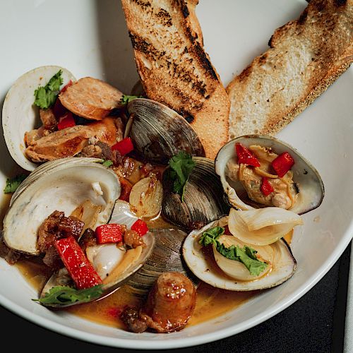 A bowl of clams topped with sausage, herbs, and red peppers, served with two pieces of grilled bread on the side.