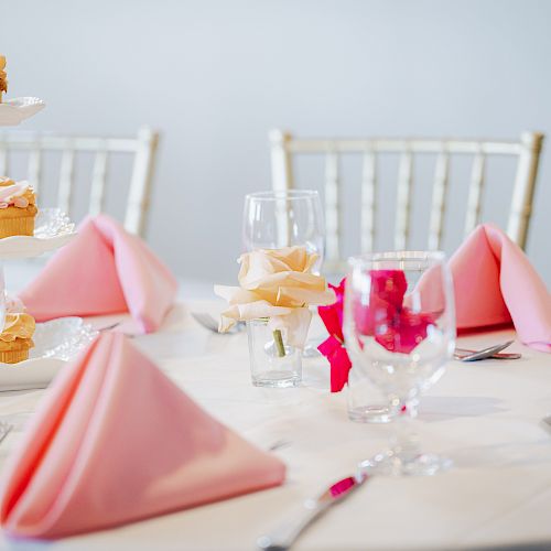 A table set for a formal event with pink napkins, glassware, silverware, a tiered stand of cupcakes, and a rose centerpiece.